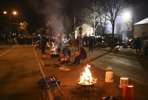 A diagonal row of campfires down a city street, people clustered around each.