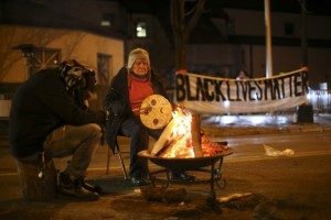 Old woman with native drum and man in shadow  by a campfire; a Black Lives Matter banner hangs in the background