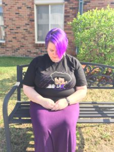 A woman with short purple hair and a black t-shirt with a cartoon of Prince sits with her head bowed.