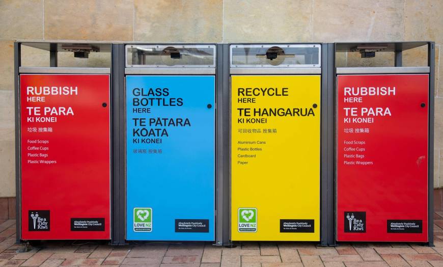 Four public trash and recycling cans, red, blue, yellow, and red. At the top they're labeled in English; beneath that in Maori; and beneath that in Chinese. 