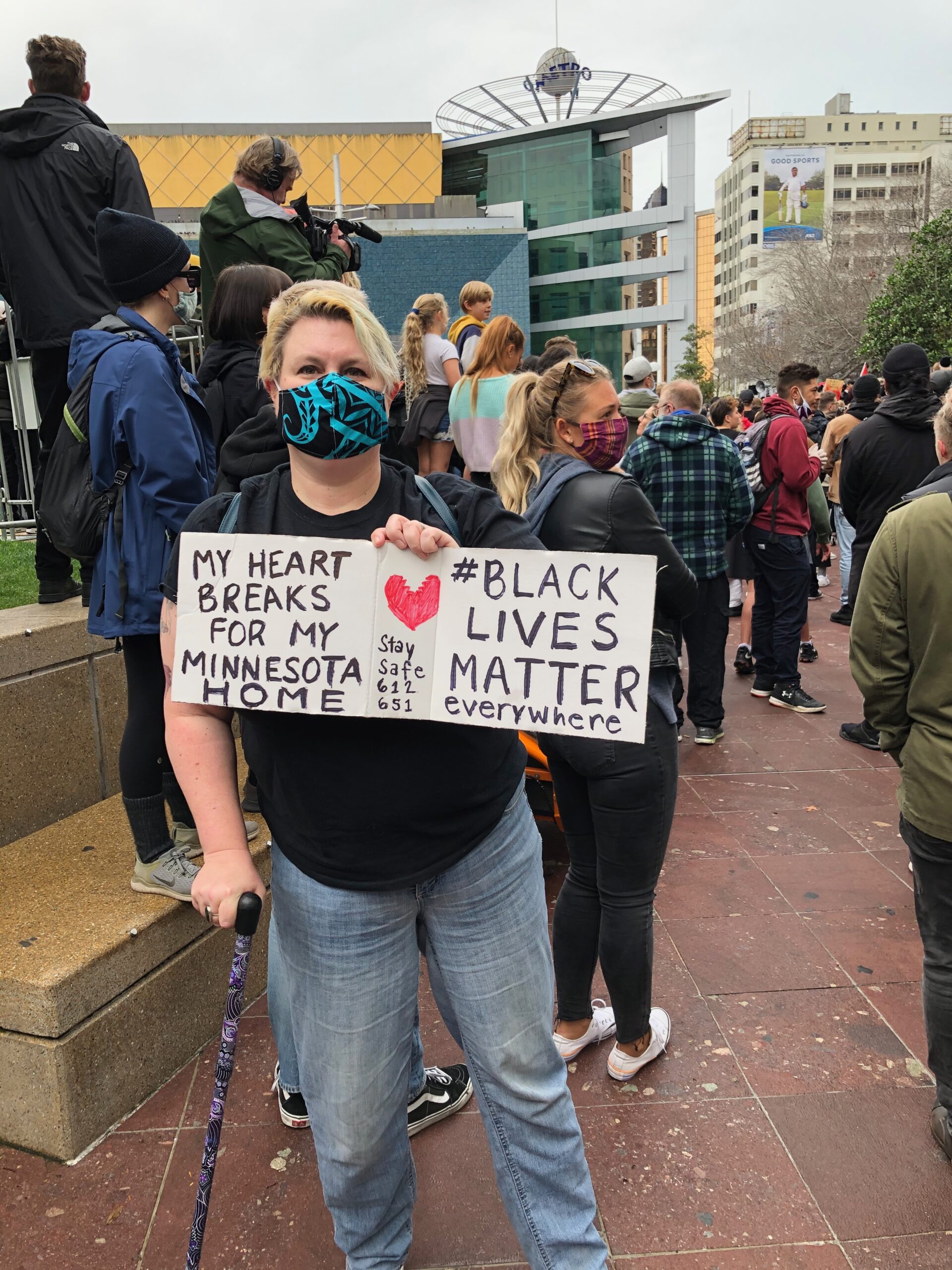 A masked-up fat white woman with short blonde hair, glasses, and a cane stands in a crowd. She holds a sign that says "My heart breaks for my Minnesota home. #BlackLivesMatter everywhere. Stay safe 651 612"