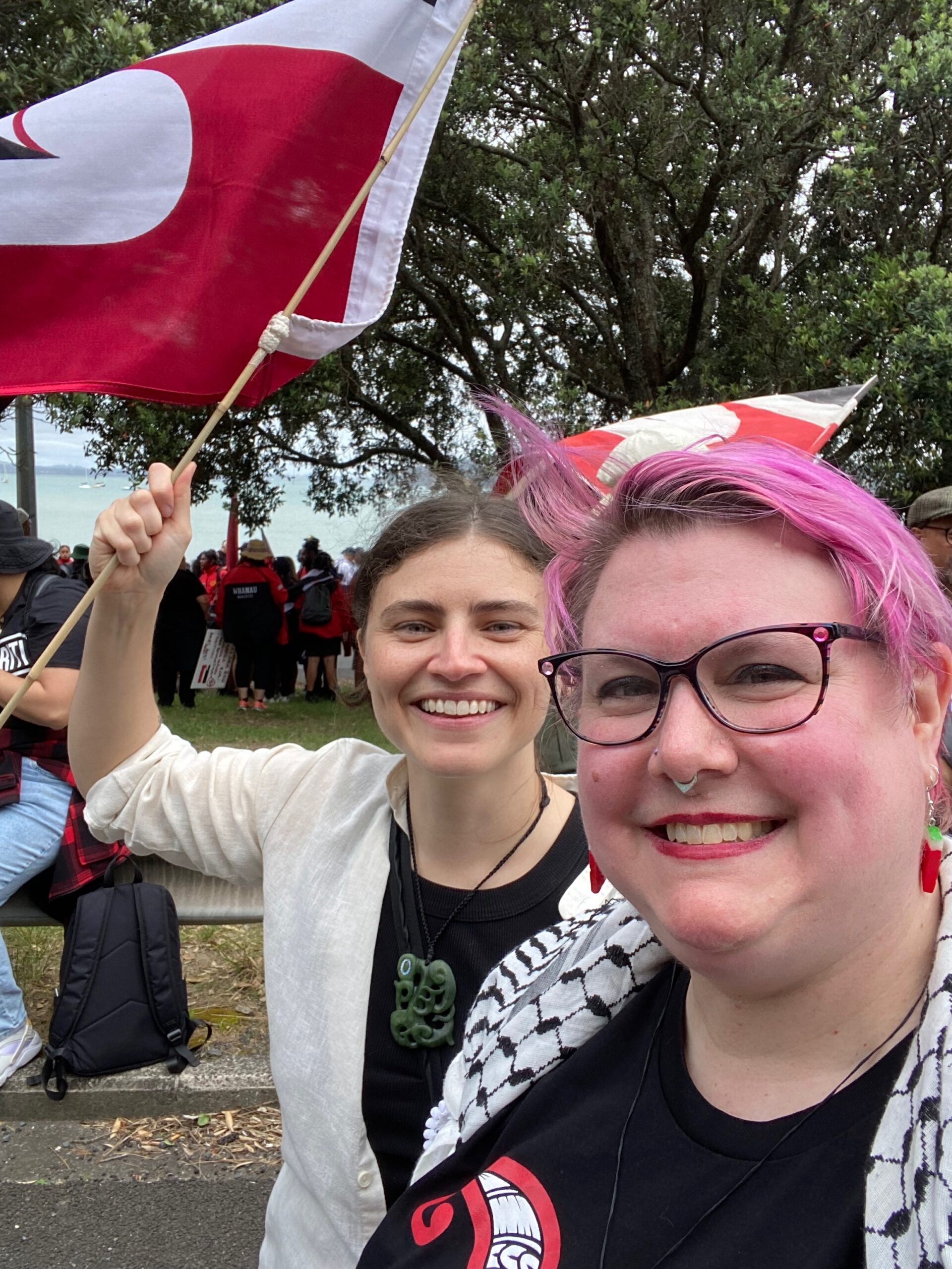 Two white women, a young one with brown hair and a fat middle aged one with short pink hair and glasses, are smiling. The young woman holds a Maori tino rangatiritanga flag.